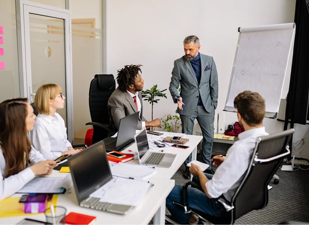 A business meeting in a modern office setting with five people discussing around a conference table