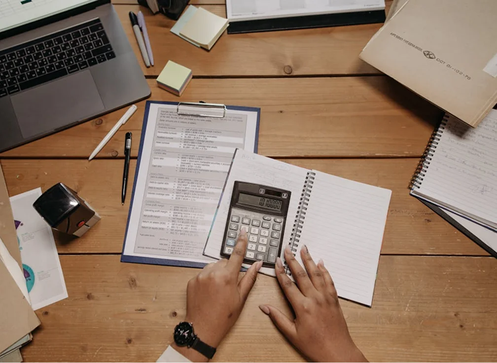A person's hands using a calculator on a wooden desk with notebooks