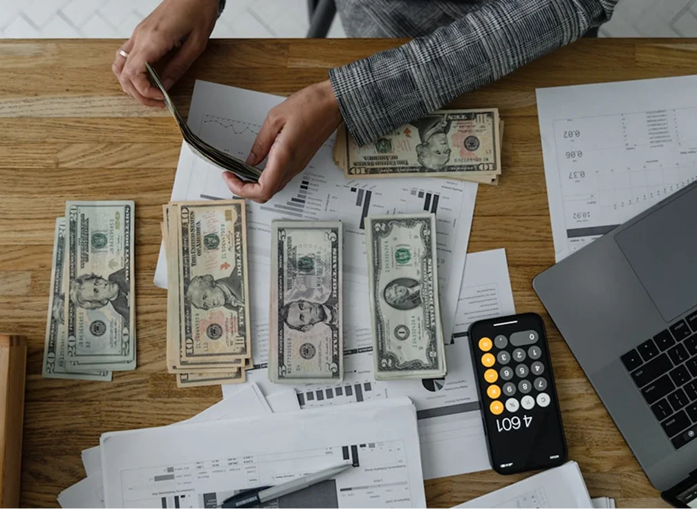 A person counting cash on a wooden table with various documents and a calculator.