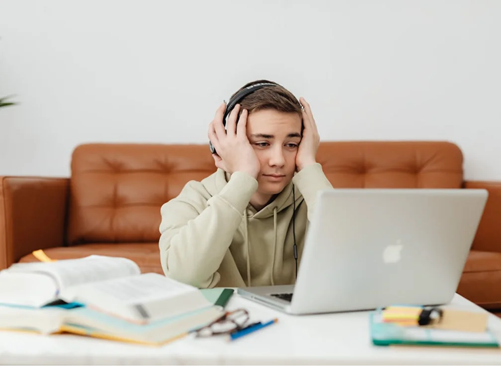 A frustrated teenage boy wearing headphones sits in front of a laptop