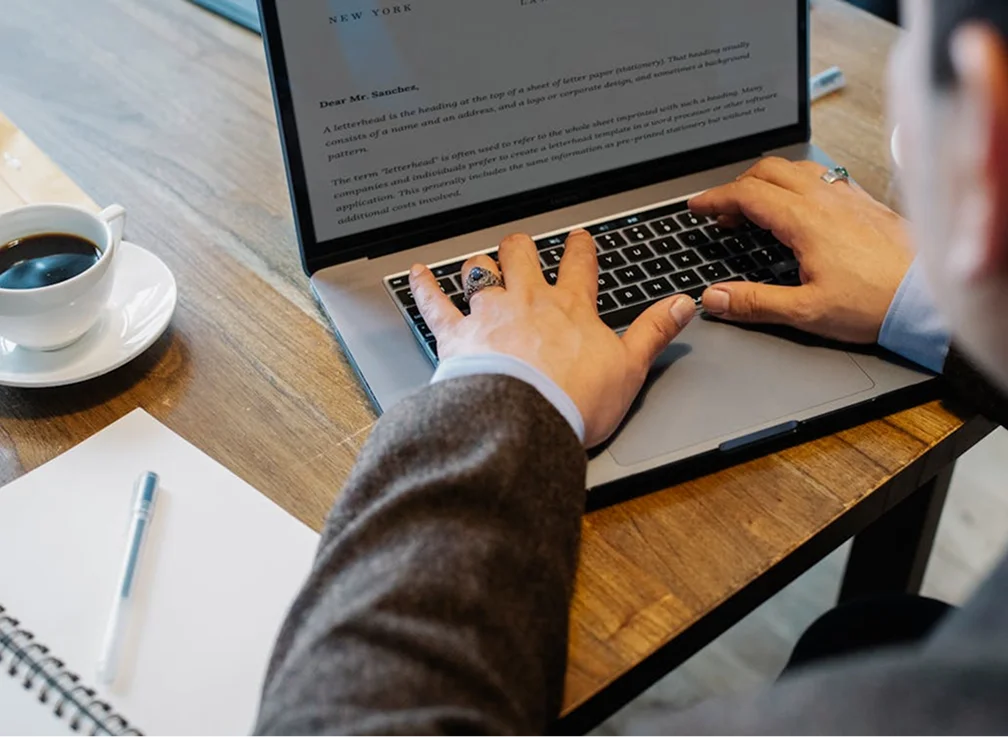 A person typing on a laptop at a wooden table with a cup of coffee and a notebook nearby.